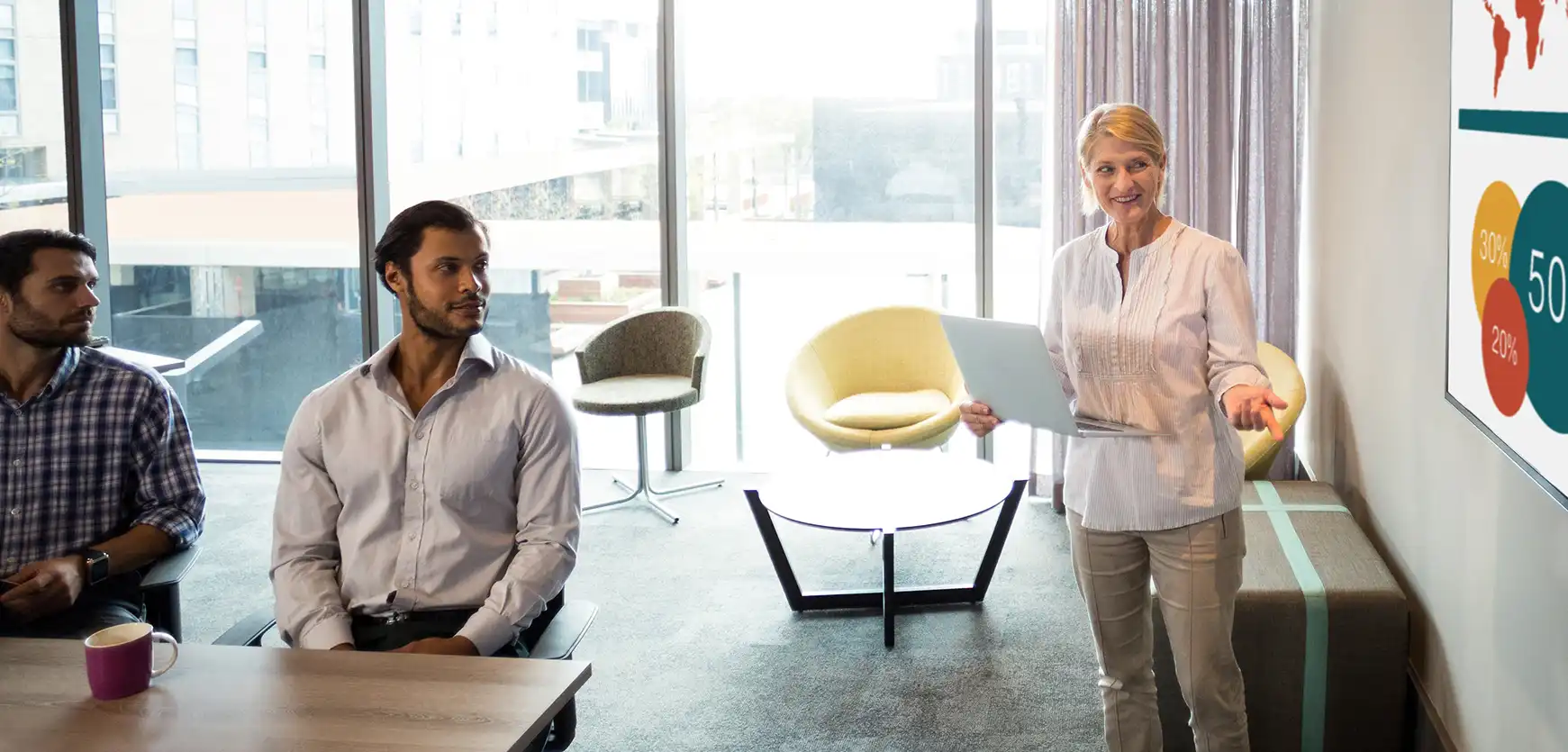A group of people in a meeting with a woman doing a presentation and pointing to the screen.